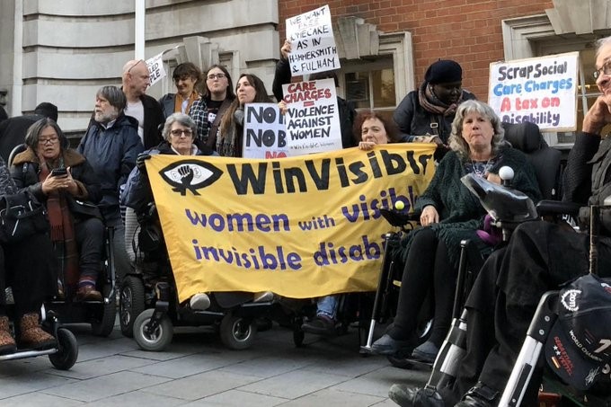 Diverse disabled protestors at Greenwich Town Hall. Women hold the WinVisible anner. Six wheelchair users in the foreground. People hold placards saying Scrap Social Care Charges, A tax on disability" and "Care Charges = Violence Against Women". We want free homecare as in Hammersmith & Fulham.