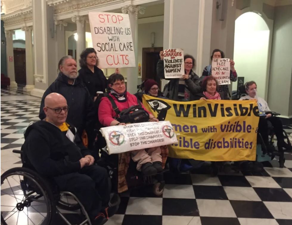 A deputation of disabled women and men, half are wheelchair users, inside Greenwich Town Hall.  Holding WinVisible and DPAC banners.  Placards say: Care charges = violence against women.  We want free homecare as in Hammersmith & Fulham.  Stop disabling us with social care cuts.  Stop the charges!