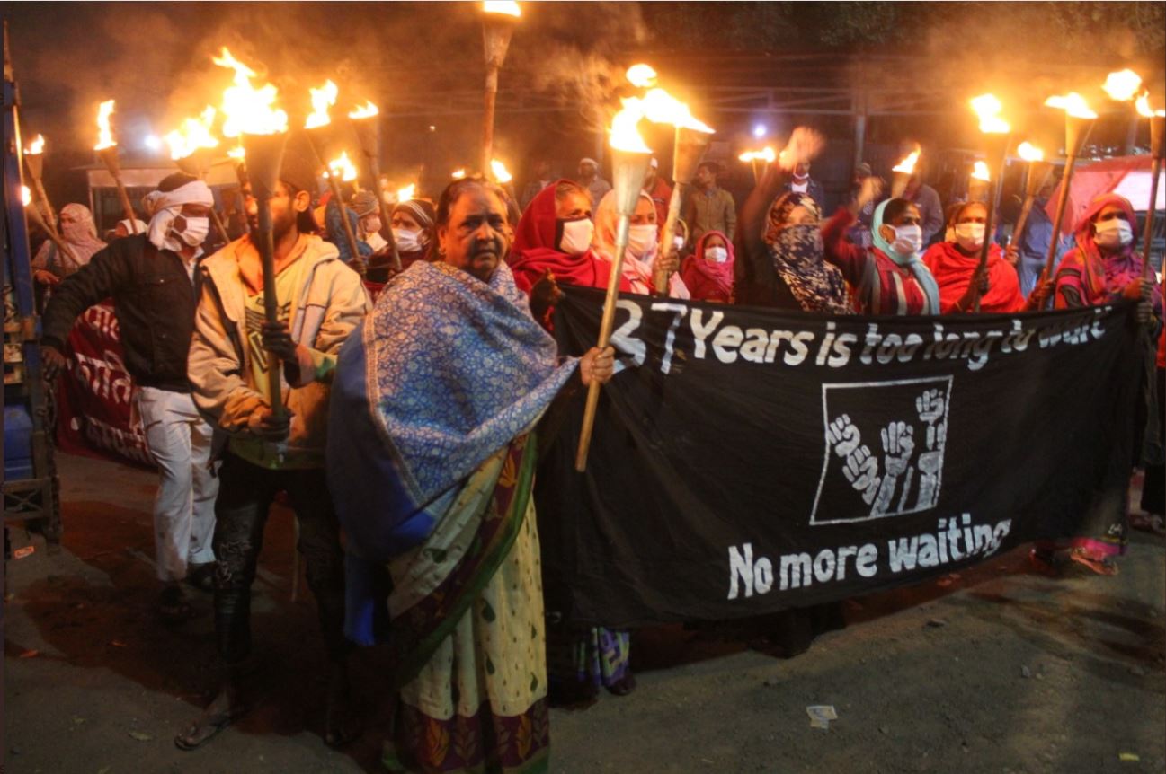 A night procession with hundreds of people. Indian women carry flaming torches and a black banner -- 37 years is too long to wait -- no more waiting. A square drawing of fists in the air.