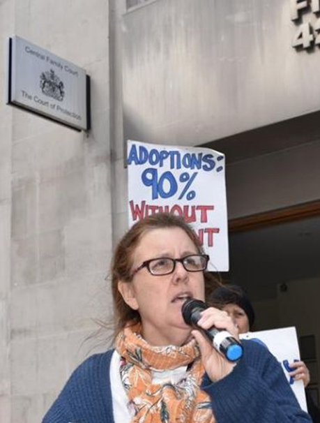 A woman speaking on the microphone outside the Central London family court. Behind her is a placard, Adoptions: 90% without consent.