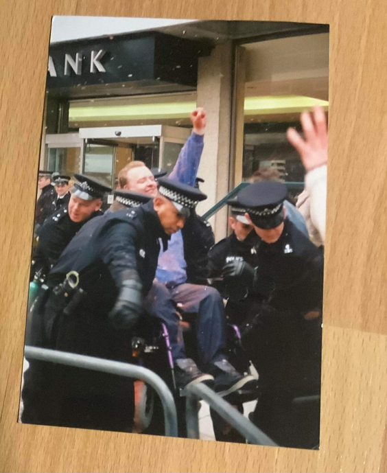 A man wheelchair user with his fist in the air -- power gesture, is carried away by the police.