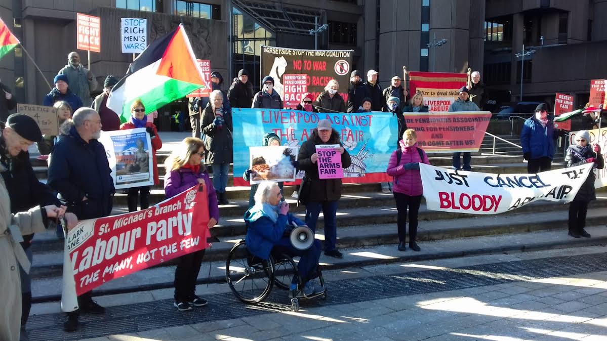 A woman wheelchair user with grey hair is speaking into a megaphone at the bottom of steps of a court building.  Around 30 supporters are on the steps holding banners, including Liverpool Against the Arms Fair, Merseyside Pensioners' Association.  Some people hold Palestinian flags.