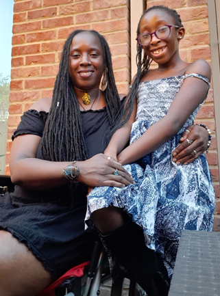 A proud disabled mum sits upright holding her daughter who is perching on the armrest of her wheelchair.  They are Afro-Caribbean and have long hair in plaits.  Both are smiling.