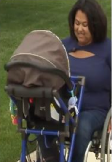 A wheelchair user mum, who is a woman of colour, looks at her baby with a smile.  The baby seat is facing her on a frame clipped to her chair.