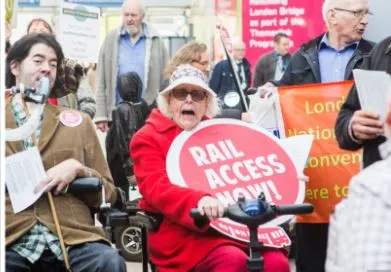 Photo of a demonstration by disabled women and men, with National Pensioners' Convention.  An older woman on a scooter holds a round placard saying Rail Access Now!  Beside her, a man wheelchair user who uses breathing support, holds a leaflet.