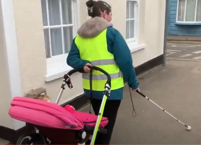 A blind mum wearing a hi-vis fluorescent vest over her jacket, is walking along using her cane.  She is pulling a buggy with her left arm behind her back.  Her child is contentedly in the buggy.