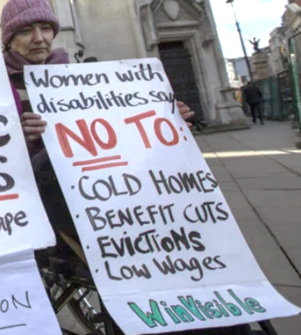 A woman wheelchair user in a woolly hat is with protesters outside the High Court.  She holds a placard: "Women with disabilities say NO TO: cold homes, benefit cuts, evictions, low wages.  WinVisible.