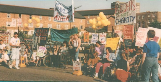 1990.  Disabled protesters, many of them women, are sitting and standing in a line with an array of colourful handmade banners and placards.  Banners include Rights Not Charity.  Not Brave Not Special but Angry.  Power not Pennies. Choices and Rights.