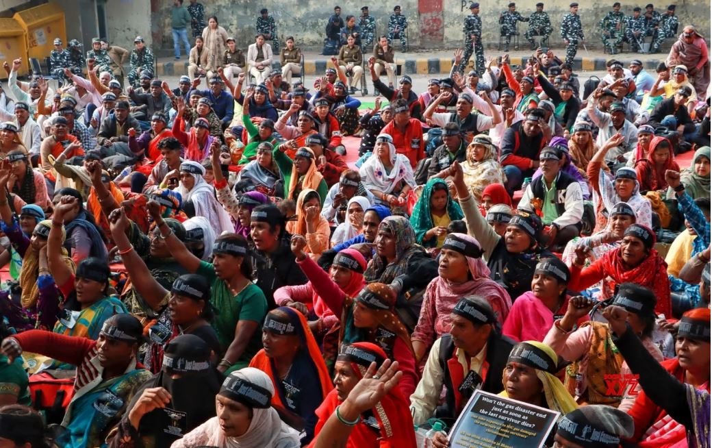 Photo shows a close-up of a protest by thousands of people.  In the photo, more than a hundred Indian women, and some men, are staging a sit-down protest outside in the road.  Many are wearing headbands with a slogan on it and have their fists in the air. One is holding a poster.  Behind them is a row of armed police.