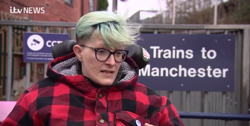 Photo of Dennis Queen at train station.  She is a wheelchair user with blonde hair and glasses.  Behind her is a sign saying Trains to Manchester.