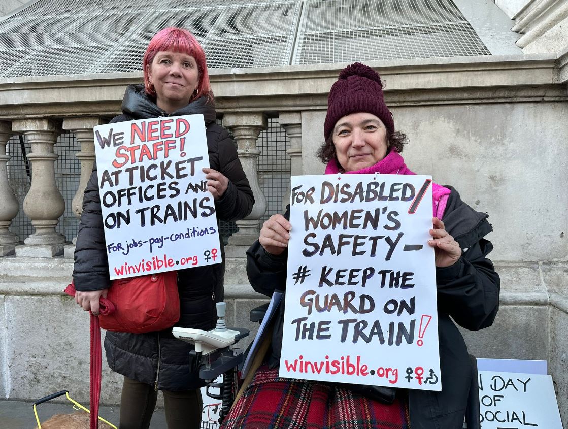 Sarah Leadbetter from National Federation of the Blind of the UK and wheelchair user Claire from WinVisible, both holding placards which read FOR DISABLED WOMEN'S SAFETY - #KEEP THE
GUARD ON THE TRAIN! winvisible.org & WE NEED
STAFF! AT TICKET OFFICES AND ON TRAINS
For jobs-pay-conditions winvisible.org. On the pavement in the street close to Number 10. Guide Dog Nellie is lying down next to Sarah and other campaign placards are seen standing up next to the wall.