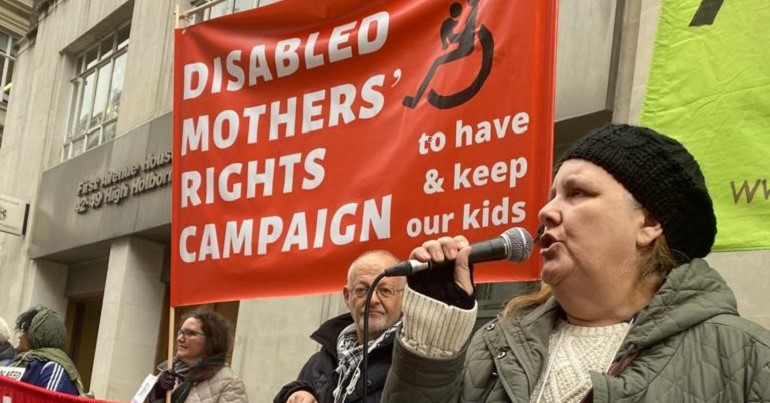 A woman in a black hat speaks into a microphone next to an orange banner with Disabled Mothers' Rights Campaign to have & keep our kids.  Outside the family court building in Holborn, London.