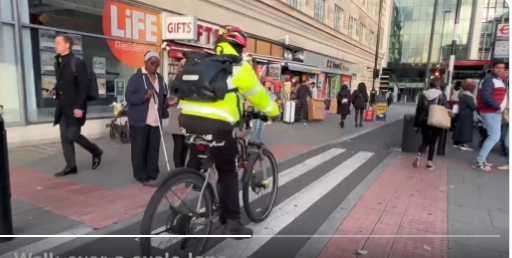 A Black woman holding her cane stands on tactile paving at the zebra crossing of a cycle lane which runs behind a bus stop.  A cyclist wearing a hi-vis jacket is riding along in front of her and has not stopped at the crossing.  