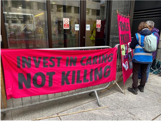 The Invest in caring not killing banner is draped on a metal crash barrier in front of Caxton House.