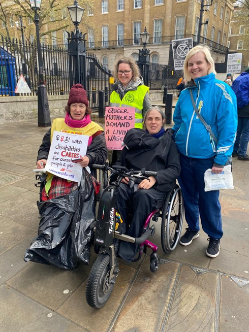 A group of four women, two wheelchair users in front. Lani's chair is made into a trike, her baby is in a sling under a waterproof cape.  Claire has a placard, women with disabilities are CARERS too! For ourselves, people & planet.  Ariane wears a hi-vis jacket and pink placard, Queer mothers demand a living wage -- Queer Strike.