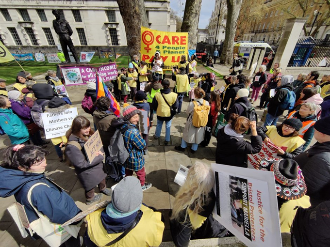 Photo from above. A crowd of mainly women, some wearing yellow tabards (sleeveless tops), listen to speakers in front of a big yellow banner which says: Women Care 4 people and planet. Pay us -- NOT billionaire polluters. Heidi Gedge holds a placard over her shoulder "Justice for the Bristol protesters -- support Mariella". Someone has a placard, Fathers join mothers vs climate chaos.