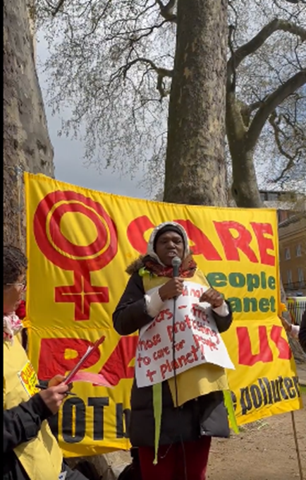 In front of the Women Care yellow banner, an African woman speaks on the microphone. She wears a placard with red writing -- Jail criminal polluters -- free those protesting who care for people and planet. Cristel from Women of Colour in the Global Women's Strike looks on. Behind are large trees.