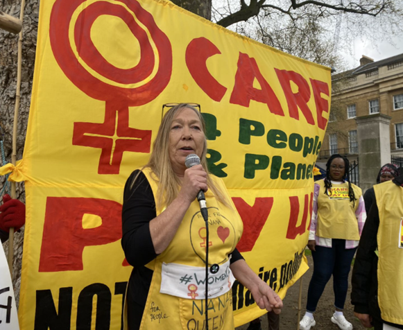A woman with blond hair speaks on the microphone in front of the Women Care banner. She is wearing a yellow tabard (sleeveless top) with a women's symbol, red heart and other writing. Women holding the banner behind her listen intently.