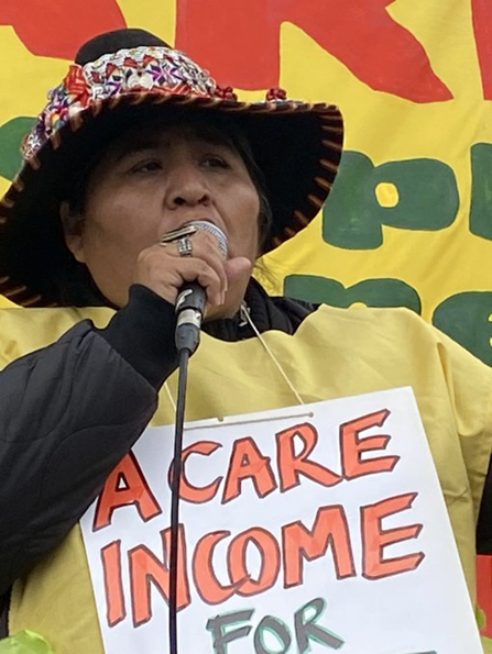 An indigenous woman from Peru speaks into the microphone. She is wearing a felt hat decorated with a multi-coloured band, yellow tabard and placard which says "A Care Income for..."