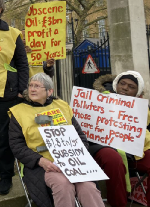 n older woman is sitting on a folding chair holding her crutch. She is wearing a yellow tabard (sleeveless top) and a placard which says Stop $1.8 trillion a year subsidy to oil, coal. She is sitting next to Trinity from the All African Women's Group. Behind them is a placard, Obscene oil: £3 billion profit a day for 50 years!