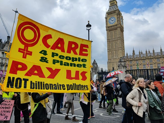 A crowd marches past Parliament and the Big Ben tower. Women carry a large yellow banner with woman's symbol and red and black lettering -- "Women Care for people & planet. Pay us -- NOT billionaire polluters." Further away are people holding umbrellas, painted with slogans like Act Now. A woman holds a pink placard, Queer mothers demand living wage.