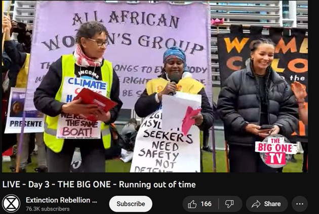 Still from The Big One TV.  Three women of colour stand in front of the Home Office building in front of banners -- All African Women's Group and Women of Colour in the Global Women's Strike.  A woman speaks on the microphone, her placard reads: LGBTQI+ asylum seekers need safety not detention.