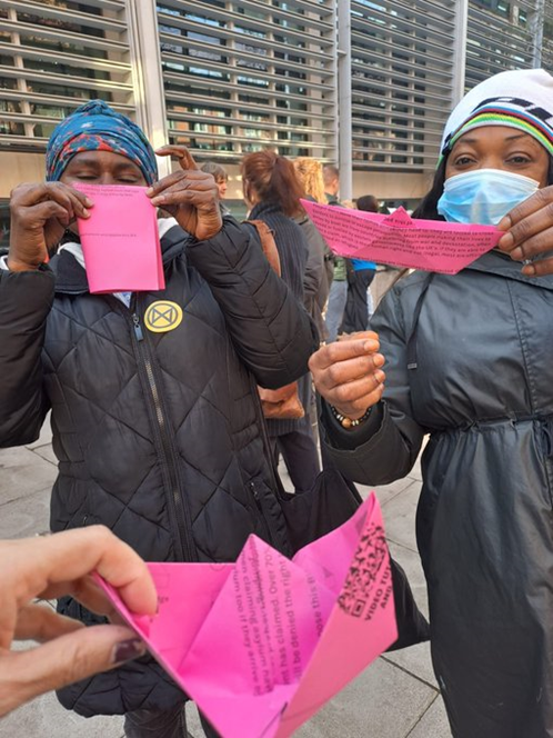 Outside the Home Office building, an African woman folds a printed piece of pink paper, while another woman with her holds a finished origami boat made from the same letter, and in the foreground another boat is held up to the camera.
