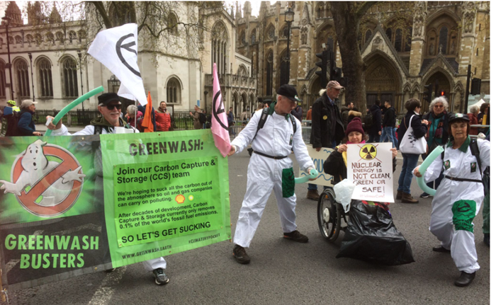 People in white overalls dressed up as ghost busters.  A man in overalls holds the Greenwash Busters banner, making fun of Carbon Capture and Storage claims.  They hold hoses up which are attached to their backpacks, and are dancing around.  Extinction Rebellion flags are attached above the green banner. A woman wheelchair user holds a placard with nuclear symbol crossed out, Nuclear power is not clean, green or safe!