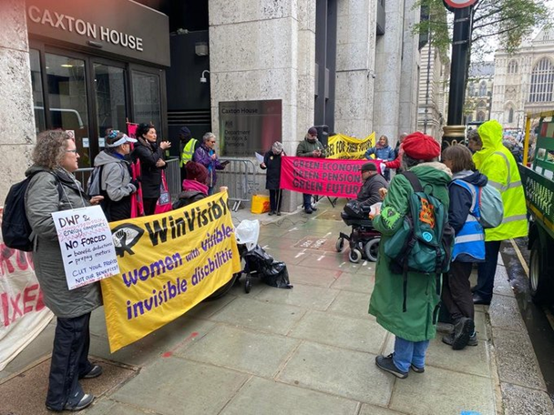 Outside a government building named Caxton House, people with banners stand in the rain. A woman is speaking on the microphone next to a sign language interpreter. Banners say WinVisible -- women with visible & invisible disabilities. Green economy -- green pensions -- green future. Ariane wears a placard "DWP and energy companies. No forced benefit deductions, prepay meters. Cut your profits, not our benefits." Security guards wear hi-vis jackets.