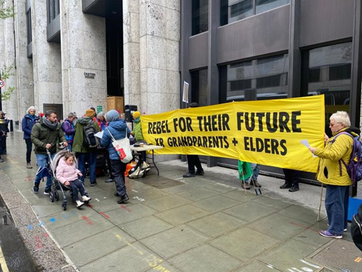 A man pushing a child in a buggy passes by the DWP building and pensioners holding a long yellow banner -- Rebel for their future -- XR grandparents + elders.