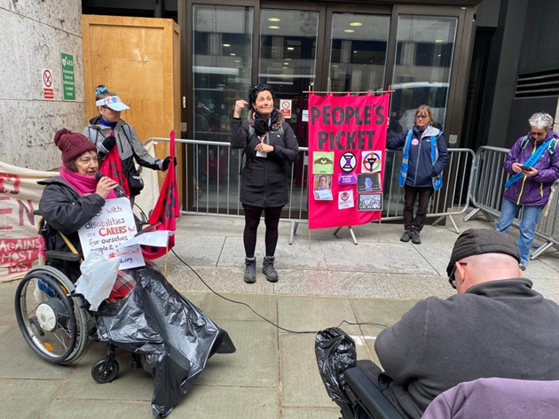 Outside a government building named Caxton House, a woman wheelchair user speaks on the microphone. A banner saying People's Picket with various photos and logos pinned to it, is placed in front of the building doors which the authorities have blocked with metal barriers. A woman interprets in sign. Another wheelchair user is there.