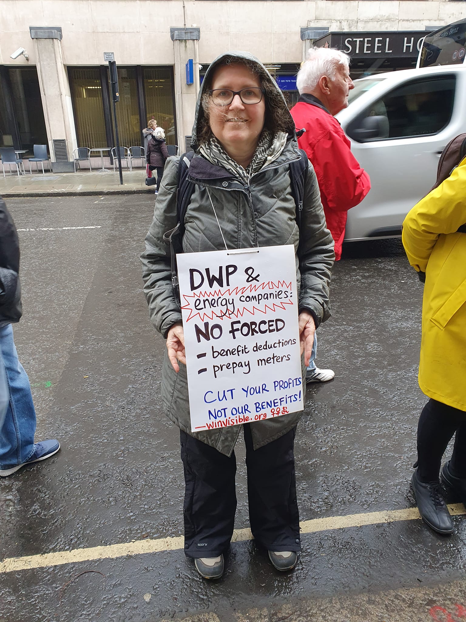 A woman wearing an anorak holds a placard: DWP & energy companies -- No forced benefit deductions/prepay meters.  Cut your profits, not our benefits! winvisible.org
