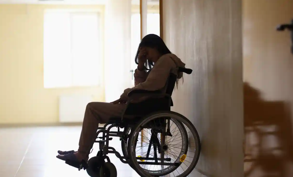 Photo of a woman wheelchair user sitting in shadow.  She is leaning her head on her hand and her face is partly hidden.  Stock photo, not a portrait.