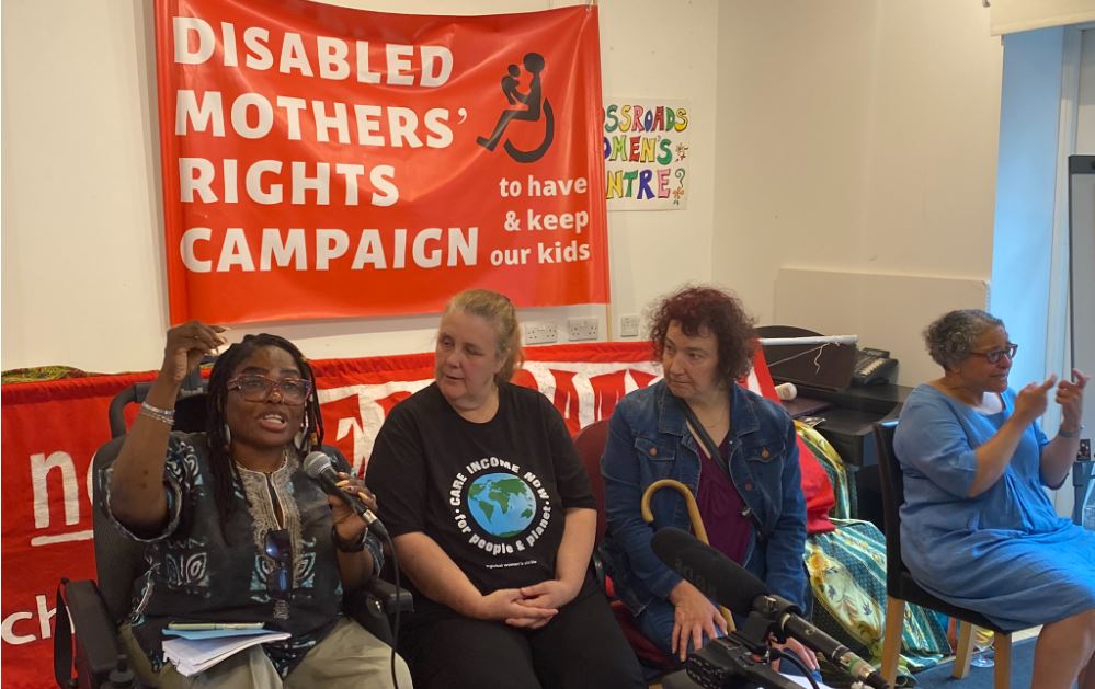 Disabled women sitting on a panel with the Disabled Mothers' Rights Campaign banner on the wall, next to the sign language interpreter. An African woman wheelchair user is speaking on the microphone gesturing with her hand.