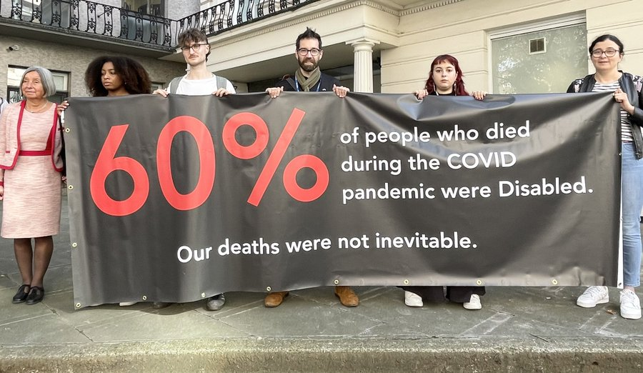 Group of disabled women and men stand behind a black banner with red and white writing: 60% of people who died during the COVID pandemic were disabled. Our deaths were not inevitable.