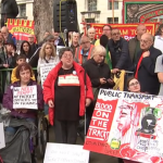 Photo of some of the big crowd with union banners behind them, listening to speakers at the RMT rally. Wheelchair users and visually impaired people are in a designated space with more room. A man has a placard resting against his legs, Save our ticket offices. A woman holds a large hand-painted board with red paint, Blood on the tracks, commemorating those who have died from train accidents. Others there have placards, humans not machines. We need staff! At ticket offices and on trains.
