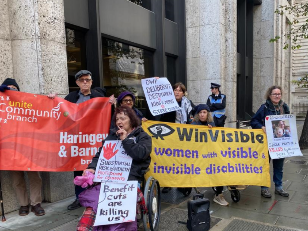 Protesters outside a large building. Women hold the WinVisible banner, next to people holding a red Unite Community Haringey & Barnet branch banner. Our placards say: "Hands off substantial risk to health, protection for claimants. Benefit cuts are killing us! DWP Deliberate Destitution of Kids and Mums is State cruelty. Remembered -- Elaine Morrall, single mum of 4, killed by UC sanction Nov 2017. " A policeman leans against the wall.