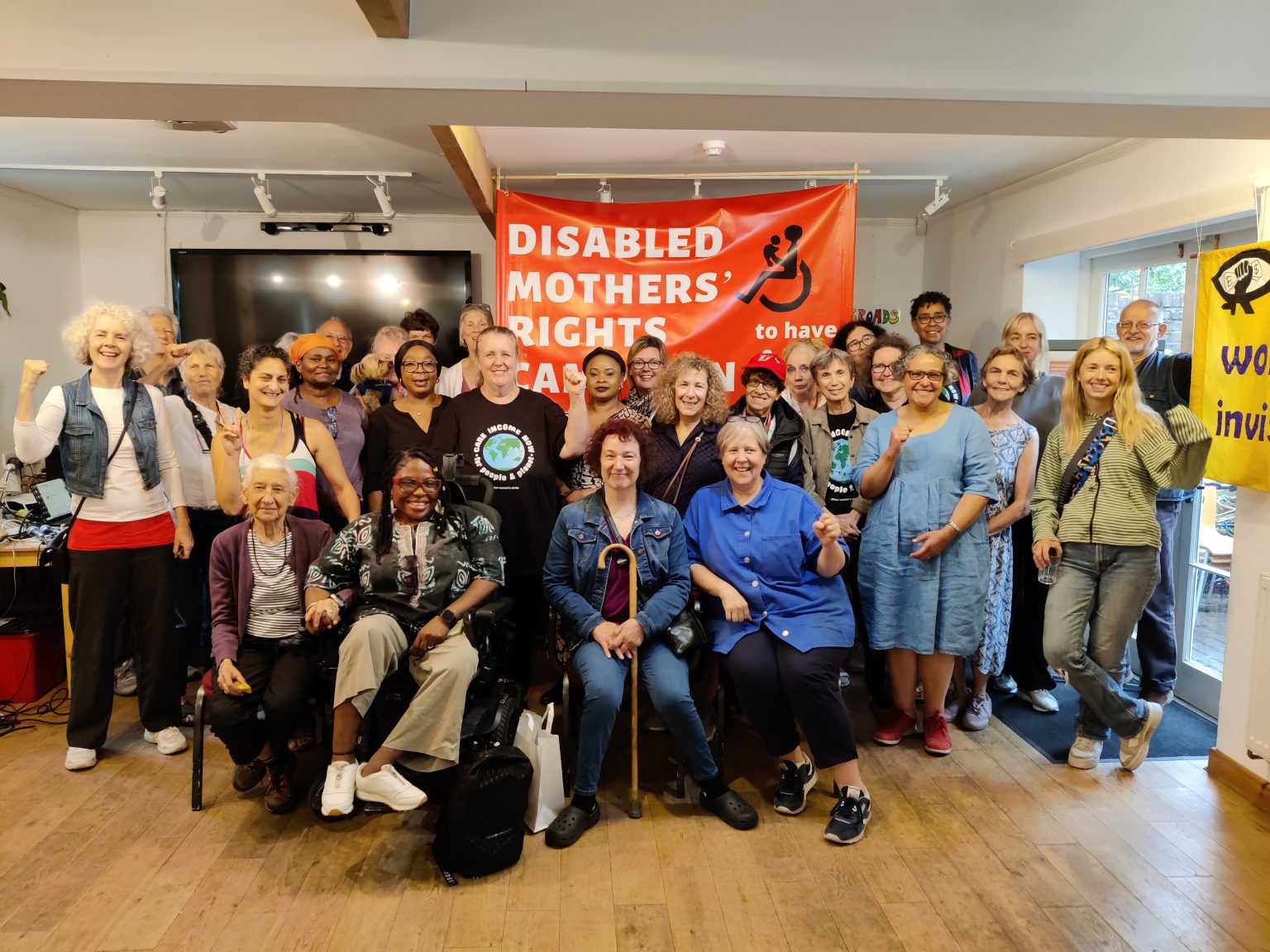 A diverse array of people, mainly women, in a meeting hall, some seated at the front, are smiling and some are making the power salute, in front of the Disabled Mothers' Rights Campaign banner.