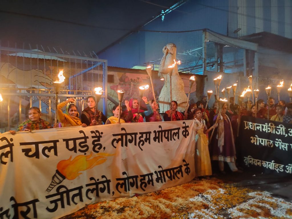 Night protest. Bhopali women hold flaming torches and two big banners.  The ground is decorated with coloured rice in writing or patterns.