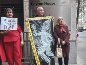 Three people stand by the entrance to Caxton House. A woman in a devil costume holds a sign, The devil loves the DWP. A man holds a cloth depicting the outline of a body with names written inside, framed by crime scene yellow tape. An older woman stands resolutely with her hands clasped.