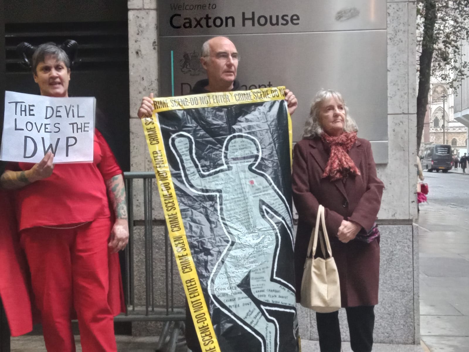 Three people stand by the entrance to Caxton House. A woman in a devil costume holds a sign, The devil loves the DWP. A man holds a cloth depicting the outline of a body with names written inside, framed by crime scene yellow tape. An older woman stands resolutely with her hands clasped.