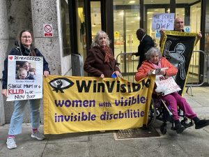 Joy Dove holds the WinVisible banner outside DWP office flanked by a woman holding a placard remembering Elaine Morrell. A woman wheelchair user holds the banner and a placard, Stop all benefit cuts and sanctions. A man holds a graphic of the outline of a body and a sign, the devil loves the DWP.