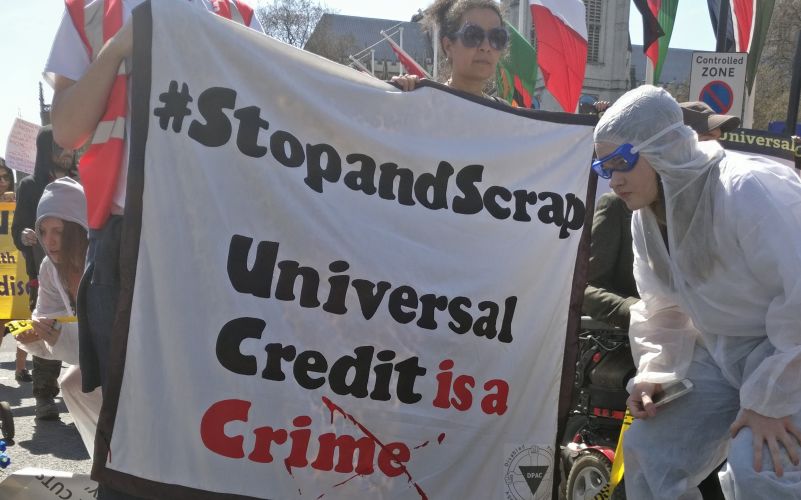 A protester in hazard clothing bends down in front of a banner a woman is holding: Stop and Scrap Universal Credit is a Crime.