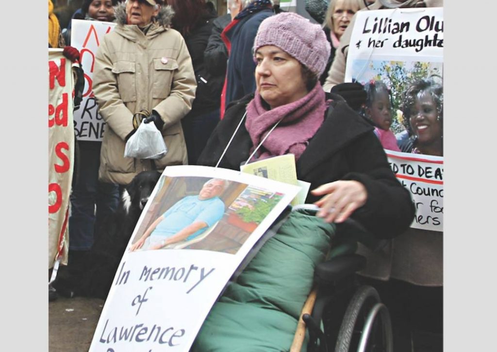 Wheelchair user woman wearing a placard, in memory of Lawrence Bond, with his photo. A crowd is behind her and a Black woman with a placard of Lillian Oluk and her daughter Lynne Matumba.