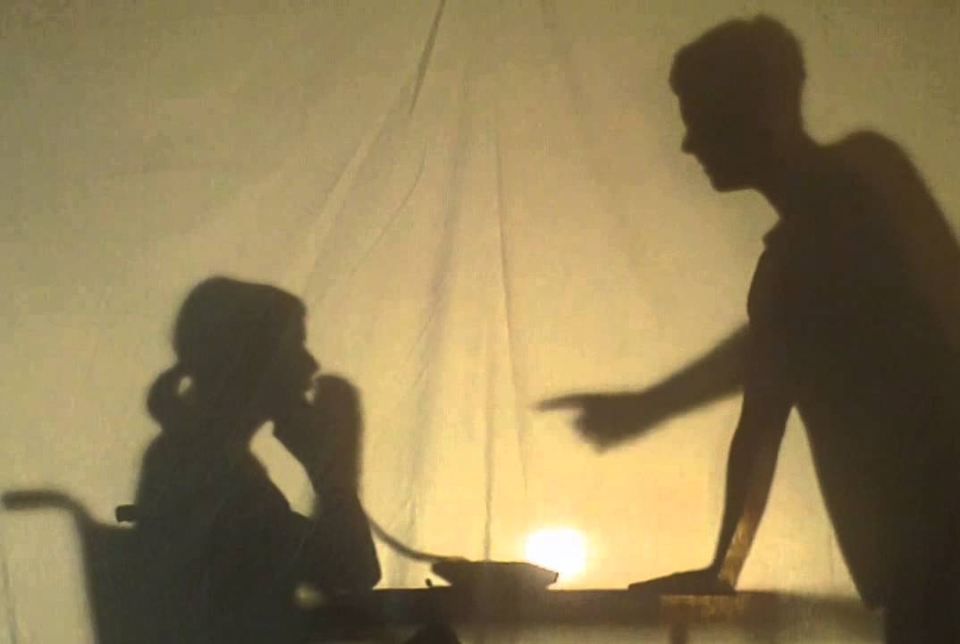 Shadows of a woman wheelchair user making a phonecall while a man stands over her, leaning towards her gesturing towards her in a threatening way.