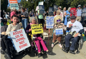 A group of three wheelchair users and a person with dwarfism at the front of the protest against the assisted dying Bill. Anna holds a placard, Assist us in living, not dying. Claire holds Invest in Caring Not Killing – Global Women’s Strike. Tamm is in drag costume and holds two placards: My life is not a priority, so why is my death? They wear a placard saying “State burden”.