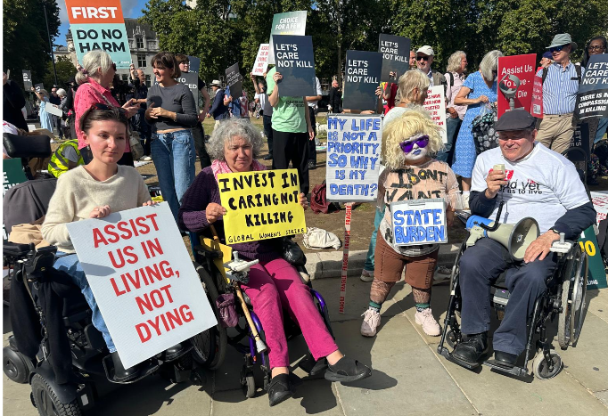 A group of three wheelchair users and a person with dwarfism at the front of the protest against the assisted dying Bill. Anna holds a placard, Assist us in living, not dying. Claire holds Invest in Caring Not Killing – Global Women’s Strike. Tamm is in drag costume and holds two placards: My life is not a priority, so why is my death? They wear a placard saying “State burden”.