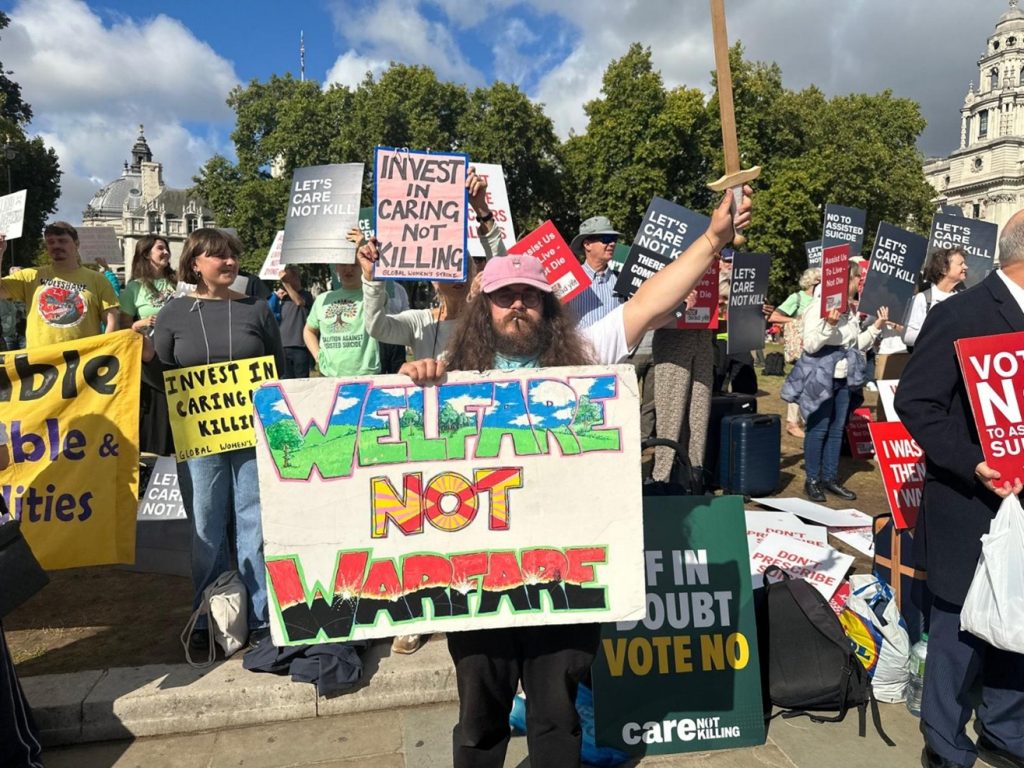 A man with a beard and long hair holds a wooden sword aloft. His placard Welfare Not Warfare has multicolour letters depicting a green Earth compared to Warfare in red and black letters depicting destroyed Earth.