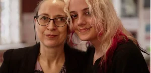 Head and shoulders portrait. A young person with long blond hair leans in to mum with glasses and brown hair. Both are smiling.