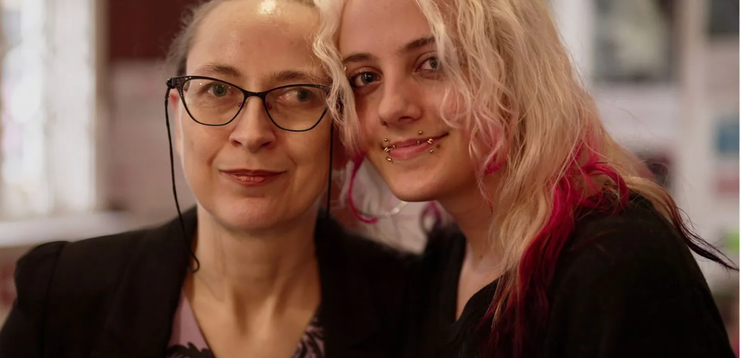 Head and shoulders portrait. A young person with long blond hair leans in to mum with glasses and brown hair. Both are smiling.
